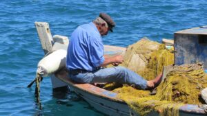 Elderly fisherman in a blue shirt mending yellow fishing nets on his wooden boat in the Mediterranean